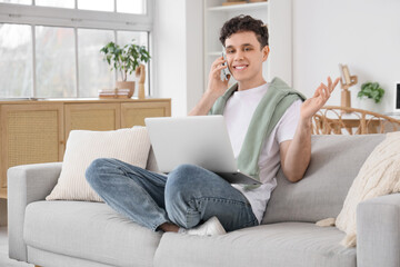 Obraz premium Young man with laptop talking by mobile phone on sofa at home