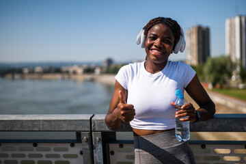 Portrait of  young sporty African woman in sportswear with headphones drinking water and showing...