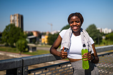 Portrait of young sporty African woman in sportswear drinking energy drink and showing thumb up...