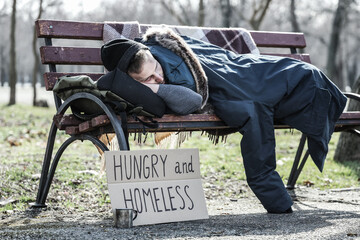 Homeless man with cardboard sleeping on bench in park