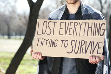 Homeless man holding cardboard with text LOST EVERYTHING TRYING TO SURVIVE in park, closeup