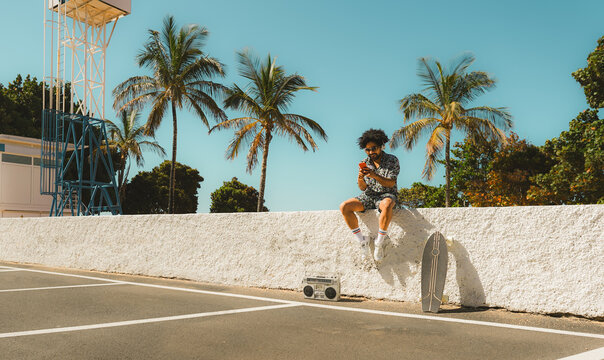 African man having fun using smartphone while listening music with beach on background - Vintage and trendy clothes concept - Focus on face