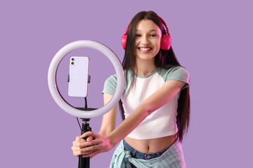 Teenage blogger in headphones recording video on lilac background