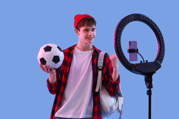 Teenage blogger with soccer ball and backpack recording video on color background