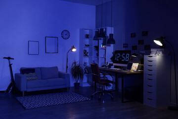 Interior of dark programmer's office with workplace, sofa and glowing lamps at night