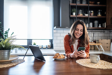 Smiling woman using smartphone and enjoying breakfast in modern kitchen