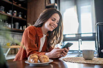 Happy woman using smartphone and having breakfast in kitchen