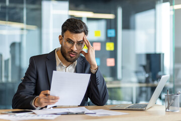 A concerned businessman in a suit is reading a document, possibly looking at financial reports or...