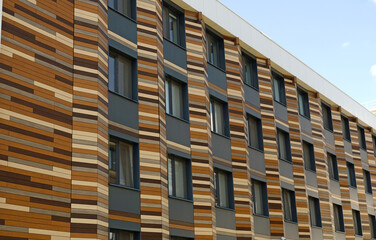 Modern building facade with distinctive striped pattern, blend of natural aesthetics and contemporary design. Dark grey sections with windows. Warm, earthy tones. Innovative architectural cladding.