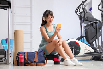 Young woman in headphones with sports bag using mobile phone at gym