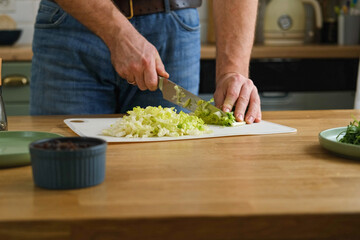 Handsome bearded man is preparing green fresh vegan salad in the kitchen at home. Male cutting...