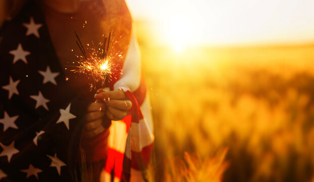 Close-up Young woman with American flag holding fireworks sparklers in a wheat field at sunset. 4th of July Celebration. Patriotic holiday, American and Flag day. Independence Day Celebration.