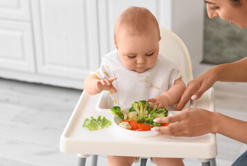 Mother feeding her baby with vegetables in high chair at home