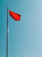 Danger sign in the form of a red flag. A red flag flutters in the wind in the blue sky by the sea during the day.