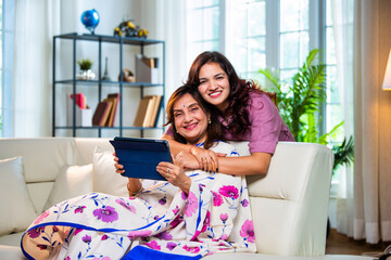 Indian mother and daughter browsing internet happily on tablet while seated in living room