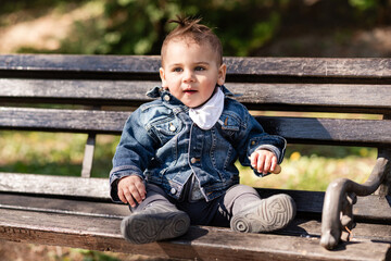 Baby boy sitting on a bench in a park wearing a denim jacket