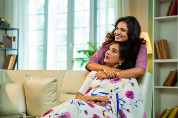 Indian daughter surprises elderly mom with a warm hug while she reads at home on sofa