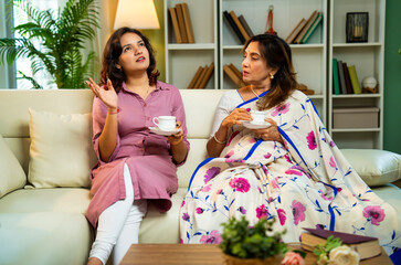 Indian senior mom and daughter happily sharing morning tea while seated on sofa at home