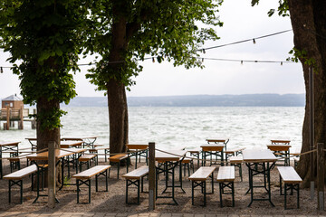 Cozy lake cafe with rustic wooden tables, benches, garlands, shaded by trees. Ideal summer relaxation spot