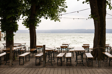 Outdoor cafe near lake, empty wooden tables shaded by big trees, garlands above. Relaxing summer mood
