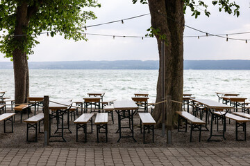 Empty wooden tables and benches of lakeside cafe under large trees with garlands, summer ambiance