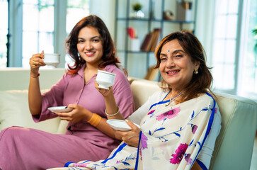 Indian senior mom and daughter happily sharing morning tea while seated on sofa at home