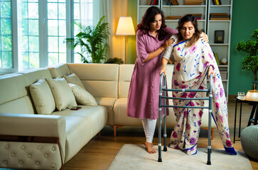 Indian daughter helping old mother walk with support due to knee pain in living room