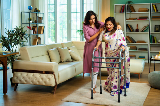 Indian daughter helping old mother walk with support due to knee pain in living room