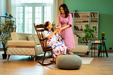 Indian woman serving orange juice to old mother in saree sitting on rocking chair at home