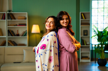 Smiling Indian mother and daughter looking at camera indoors with positive expressions
