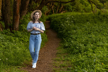 A woman in a hat and jeans walks along a path in the forest.