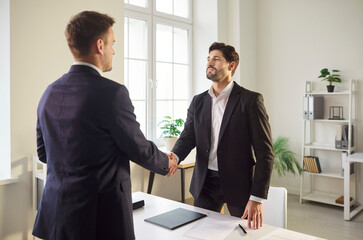 Confident man shaking hands with his partner standing in office celebrating success or making a good deal signing a contract. Young cheerful male company employees reaching agreement on meeting.