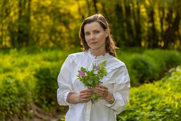 a woman in a white shirt with ruffles, holding a bouquet of green leaves and flowers. It stands...