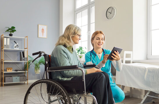 Young friendly female nurse or social worker using modern tablet with senior woman with disability in wheelchair. Caregiver learning her elderly patient using digital gadget in nursing home.