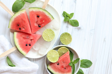 Ripe watermelon pieces on sticks like ice cream. Watermelon, lime and mint. Summer fruits background. Flat lay. Watermelon slices in white plate top view, wooden table and lime halves. Summer food