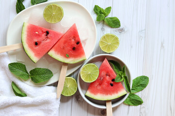 Ripe watermelon pieces on sticks like ice cream. Watermelon, lime and mint. Summer fruits background. Flat lay. Watermelon slices in white plate top view, wooden table and lime halves. Summer food