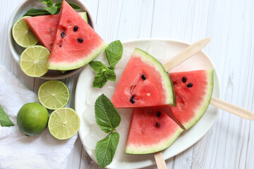 Ripe watermelon pieces on sticks like ice cream. Watermelon, lime and mint. Summer fruits background. Flat lay. Watermelon slices in white plate top view, wooden table and lime halves. Summer food