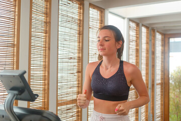 A young woman is jogging on a treadmill in a well-lit fitness area with bamboo shades, focusing on...