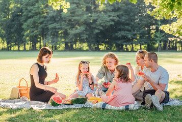 Big family sitting on the picnic blanket in city park during weekend Sunday sunny day. They are...