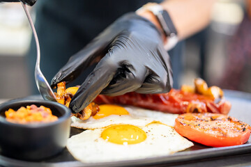Close-up of a chef plating a gourmet breakfast with fried eggs, grilled tomato, mushrooms, and salsa. Professional food styling and culinary presentation in a kitchen setting.