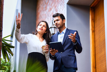 Indian coworkers in formalwear consult outdoors with tablet and coffee, standing near modern office