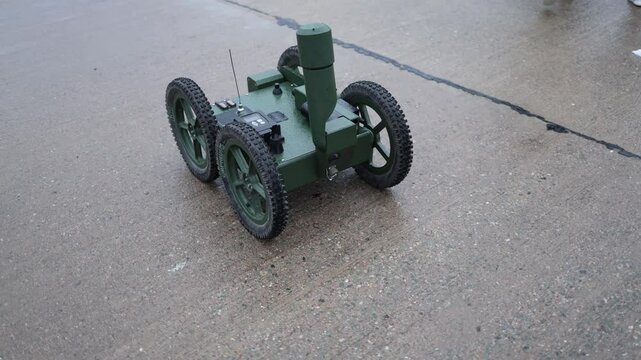 Military ground robot stands idle on wet concrete with rugged wheels sensors and antenna ready for reconnaissance missions under light rain and gray urban atmosphere