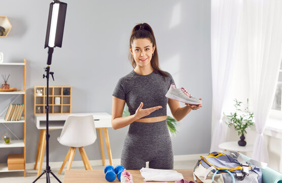 Female blogger is showing sports shoes and equipment, preparing to create content about workout. Woman presents sneakers to her online followers in home studio with professional lighting equipment.
