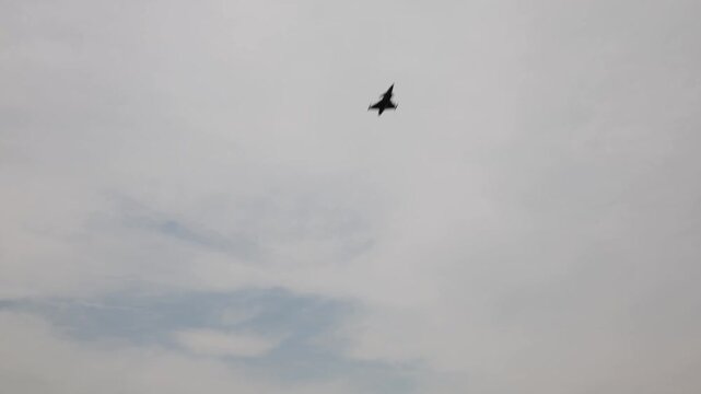 Fighter jet tilts mid-turn under pale overcast sky with soft blue patches breaking through clouds blending speed grace and sharp control into a quiet yet powerful aerial moment