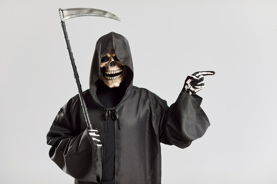 Portrait of creepy man in image of death with scythe pointing his finger to side. Man in creepy skull mask and black hooded robe points to place for text. Day of dead and Halloween concept.