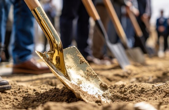 Golden shovel pushes into soil at groundbreaking ceremony other shovels and attendees in background
