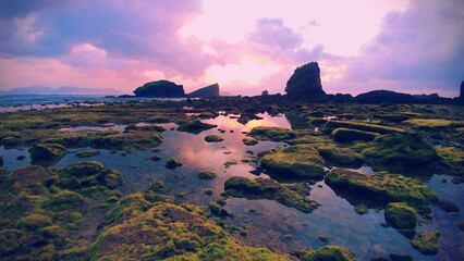Wide-angle shot of a vibrant sunrise over Jember’s rocky coast, with pink-orange skies reflecting in calm tide pools creating a majestic and ethereal coastal landscape at daybreak