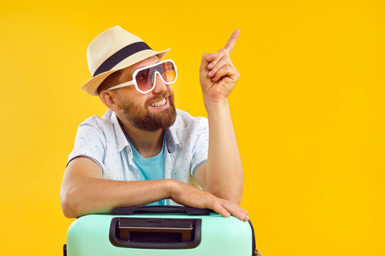 Portrait of young happy funny man tourist in sunglasses and beach hat with suitcase pointing index finger to the side on yellow background. Vacation advertising, summer holiday and travel concept.