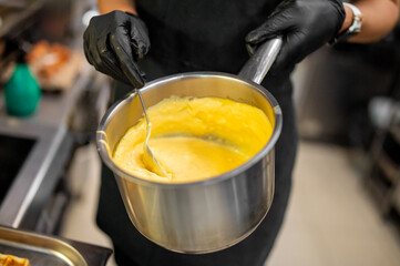 Chef in black gloves stirring creamy yellow sauce in metal saucepan on stove, highlighting...