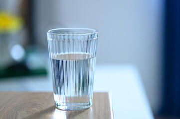 Glass containing clear water sits on a smooth wooden table in a well-lit indoor environment,...